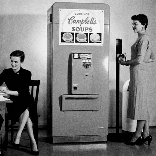 Women in break room with one purchasing a can of soup from a vending machine.